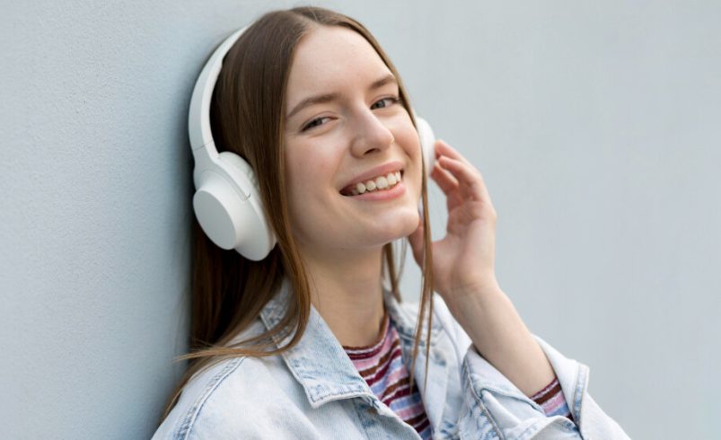 A woman wearing headphones, leaning against a wall, enjoying music. She may be using YouTube to MP3 converters.